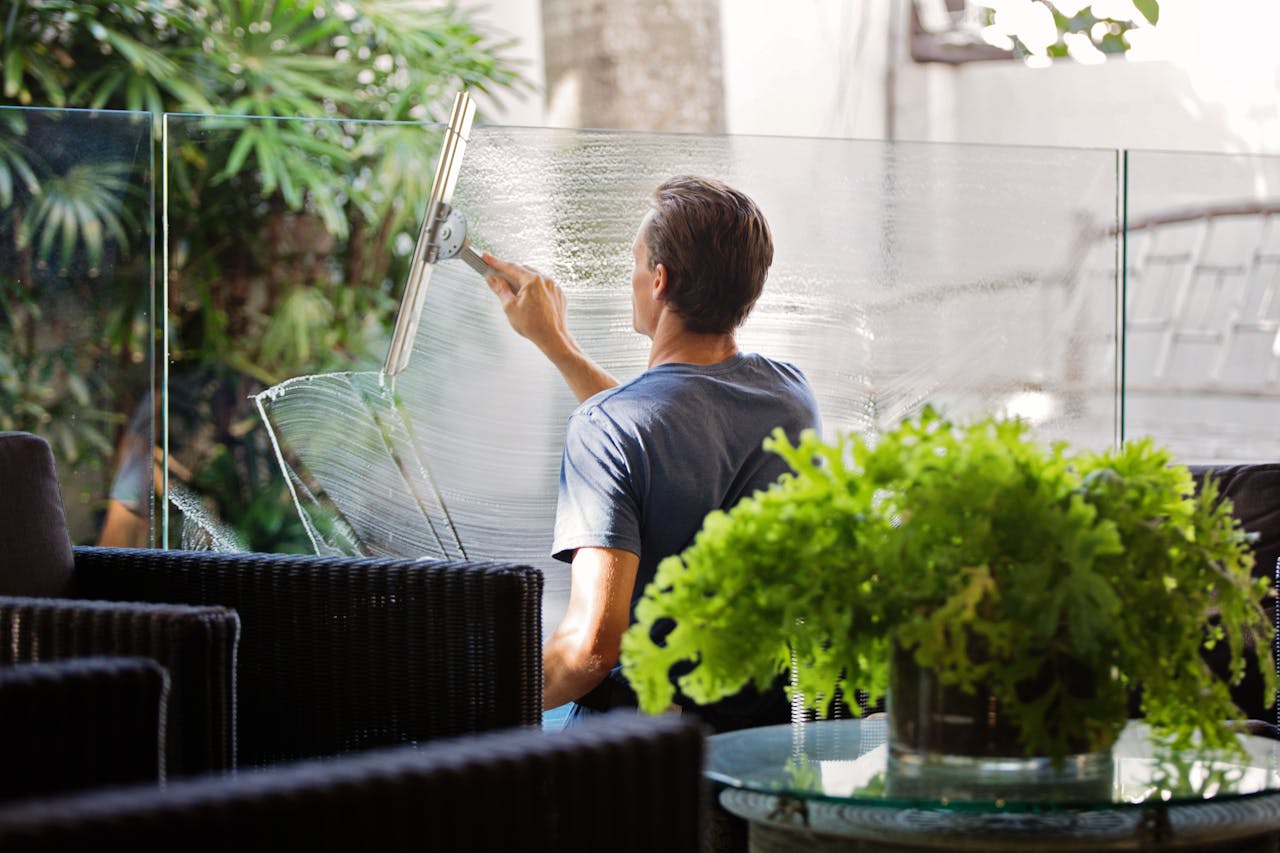 heros-img A man cleaning a glass barrier outdoors with a squeegee, surrounded by greenery.